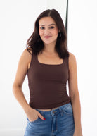A woman with dark brown hair wearing a fitted brown tank top and blue jeans, standing against a bright white background.