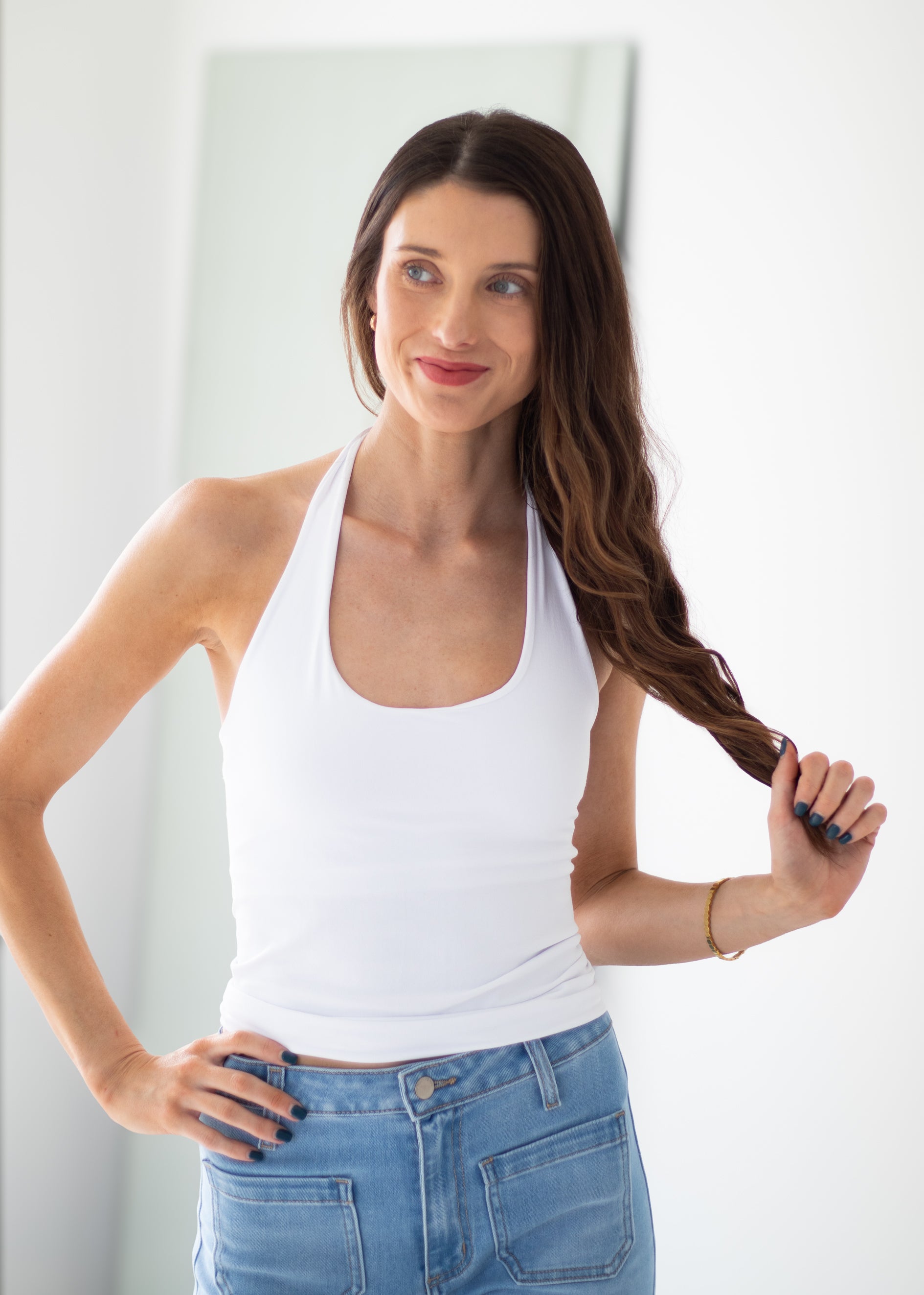 A woman with long brown hair wearing a white halter tank top and blue jeans, holding a section of her hair in a bright indoor setting.