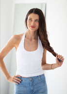 A woman with long brown hair wearing a white halter tank top and blue jeans, holding a section of her hair in a bright indoor setting.