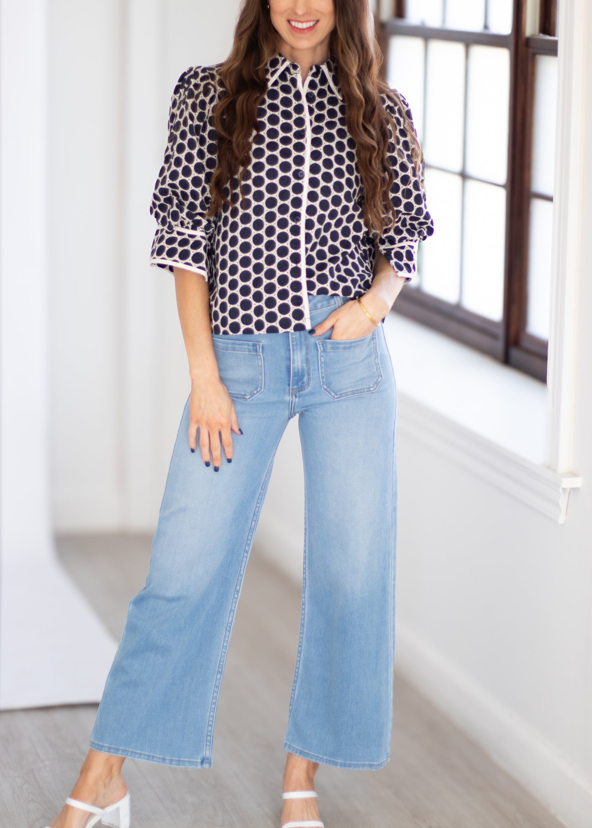 A woman wearing a black and white polka dot blouse with three-quarter sleeves and light blue wide-leg cropped jeans, standing in a bright room with a window.