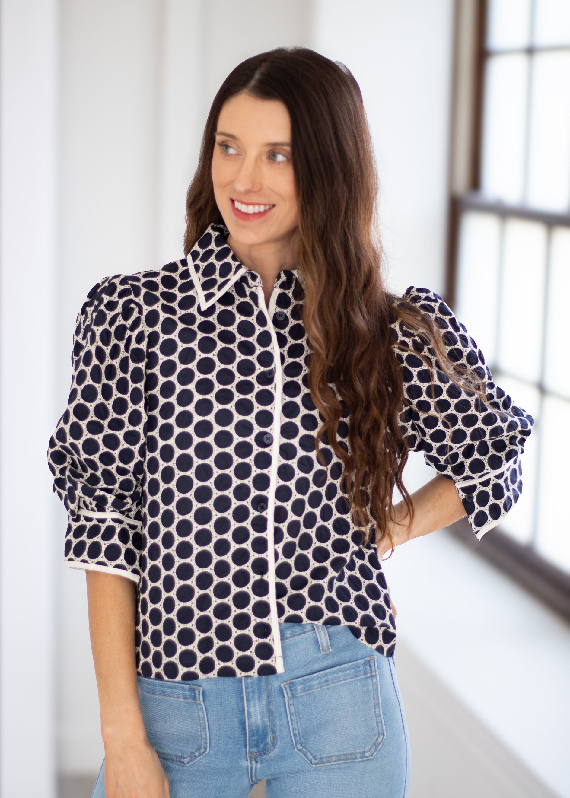 A woman wearing a black and white polka dot blouse with puffed sleeves and light blue jeans, standing in a bright indoor setting.