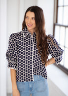 A woman wearing a black and white polka dot blouse with puffed sleeves and light blue jeans, standing in a bright indoor setting.