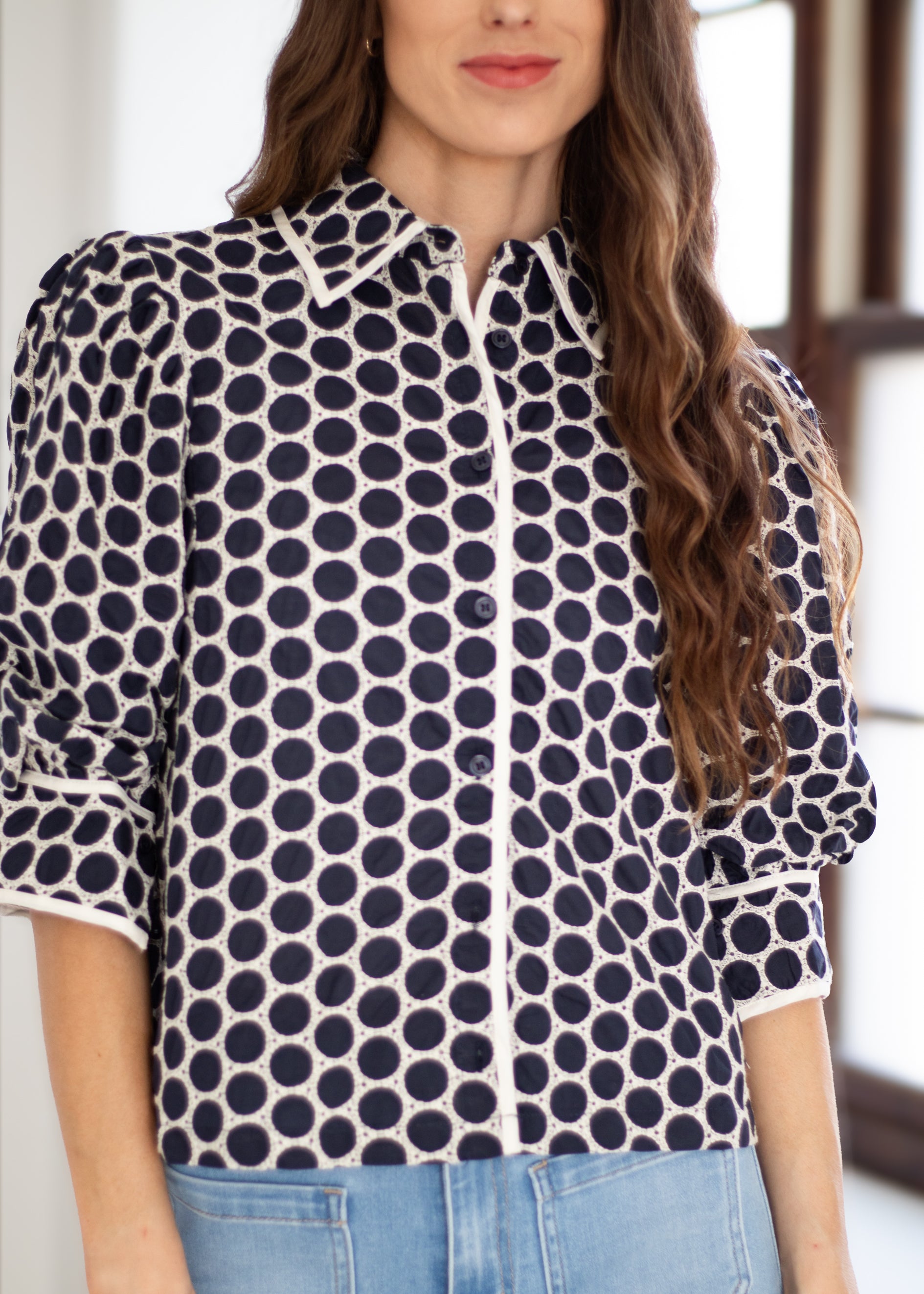 A woman wearing a navy and white polka dot button-up blouse with puff sleeves, paired with light blue jeans.