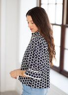 A woman with long brown hair is shown in profile wearing a navy and white patterned long-sleeve top with light blue jeans, standing in a brightly lit room with a window in the background.