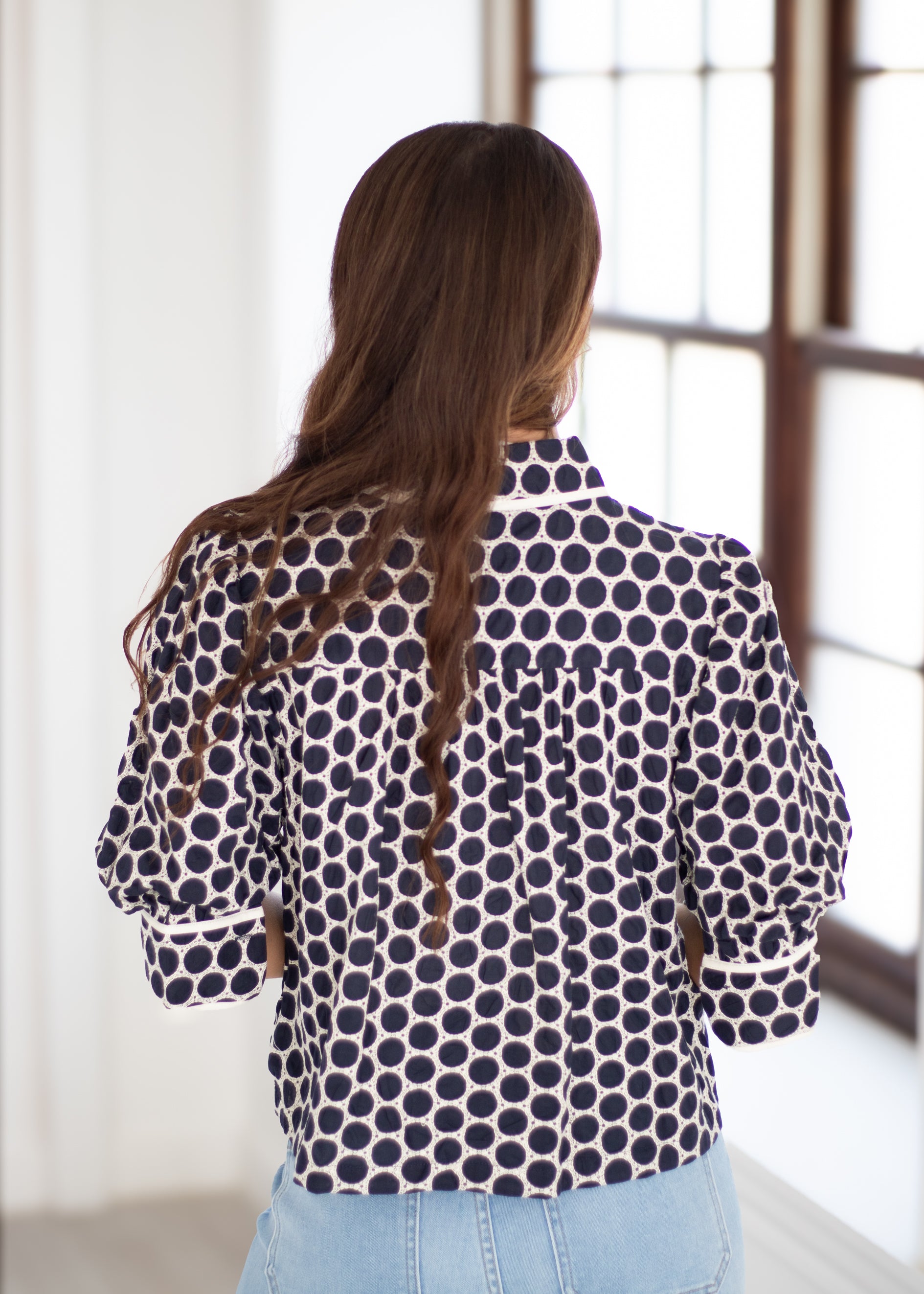 A woman viewed from behind wearing a navy blue and white polka dot patterned blouse with light blue jeans, standing in a brightly lit room with a window.