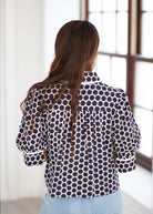 A woman viewed from behind wearing a navy blue and white polka dot patterned blouse with light blue jeans, standing in a brightly lit room with a window.