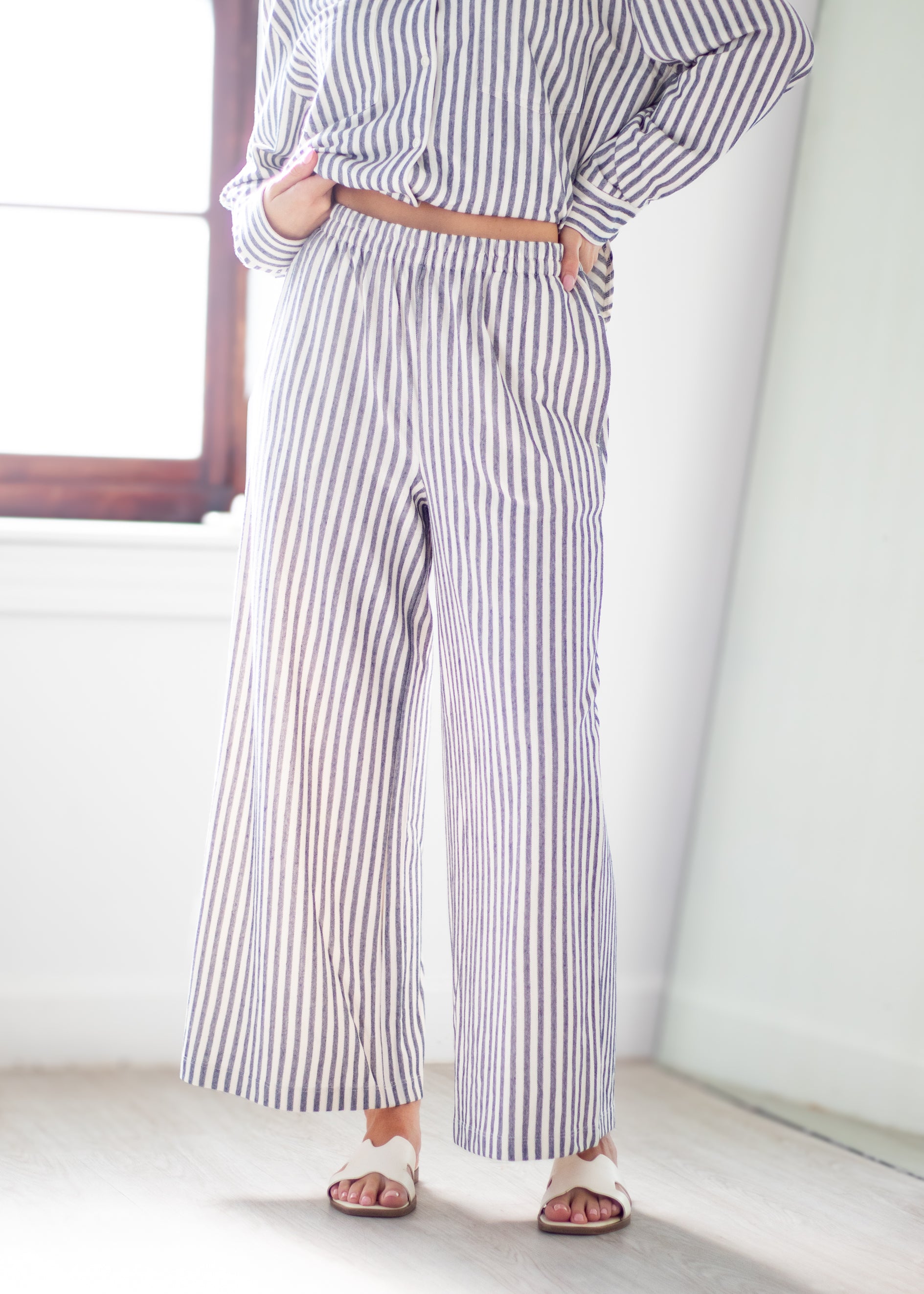 A person wearing a matching long-sleeve button-up shirt and wide-leg trousers set with vertical navy and white stripes, styled with a tan belt and white slide sandals in a bright indoor setting.