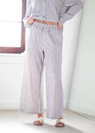 A person wearing a matching long-sleeve button-up shirt and wide-leg trousers set with vertical navy and white stripes, styled with a tan belt and white slide sandals in a bright indoor setting.