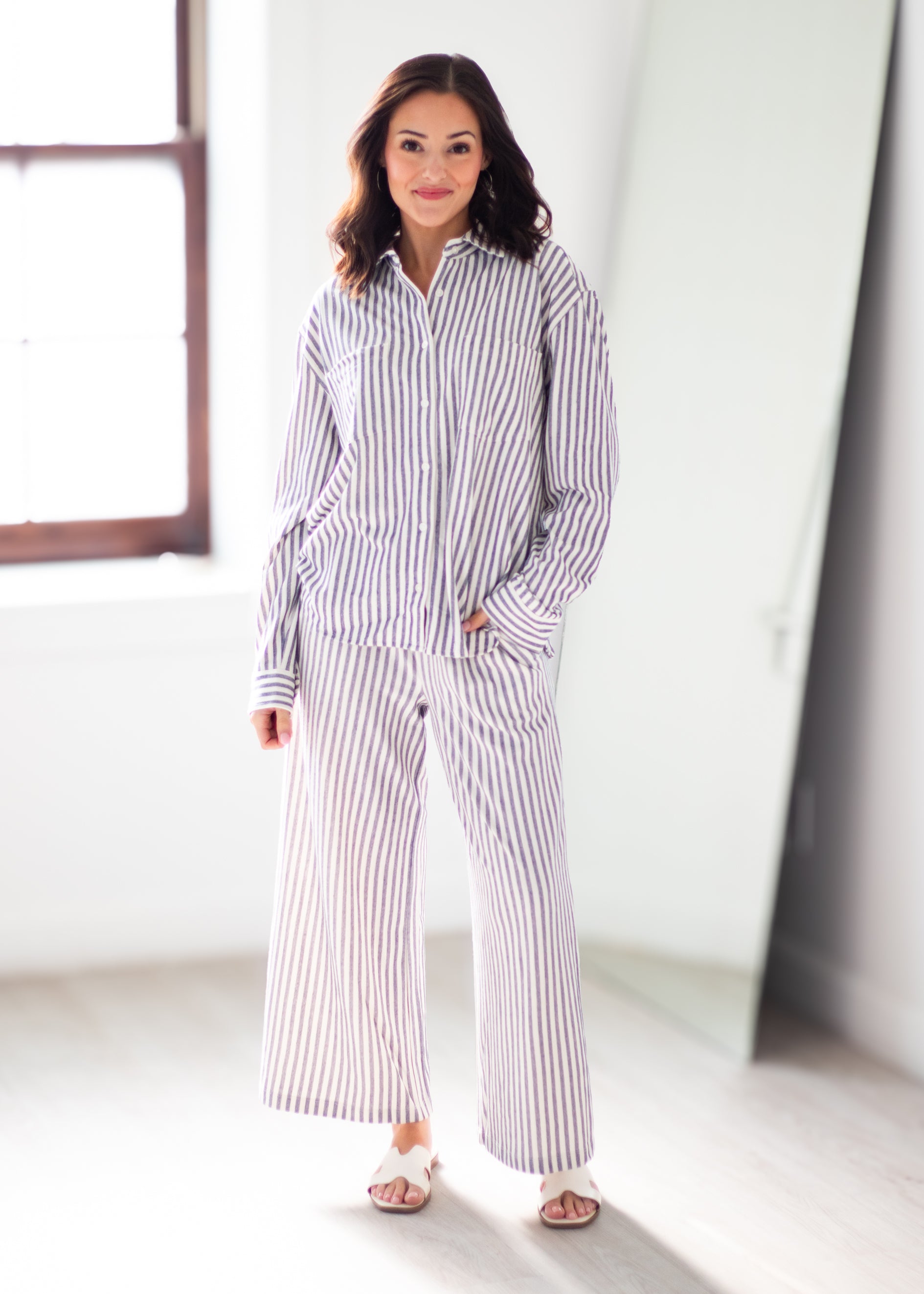 A woman wearing a matching two-piece pajama set with vertical stripes, standing in a bright, modern room with a window and mirror.