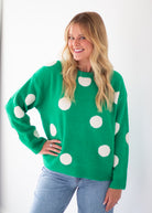 A woman wearing a bright green knit sweater with large white polka dots, paired with light blue jeans, standing against a plain white background.