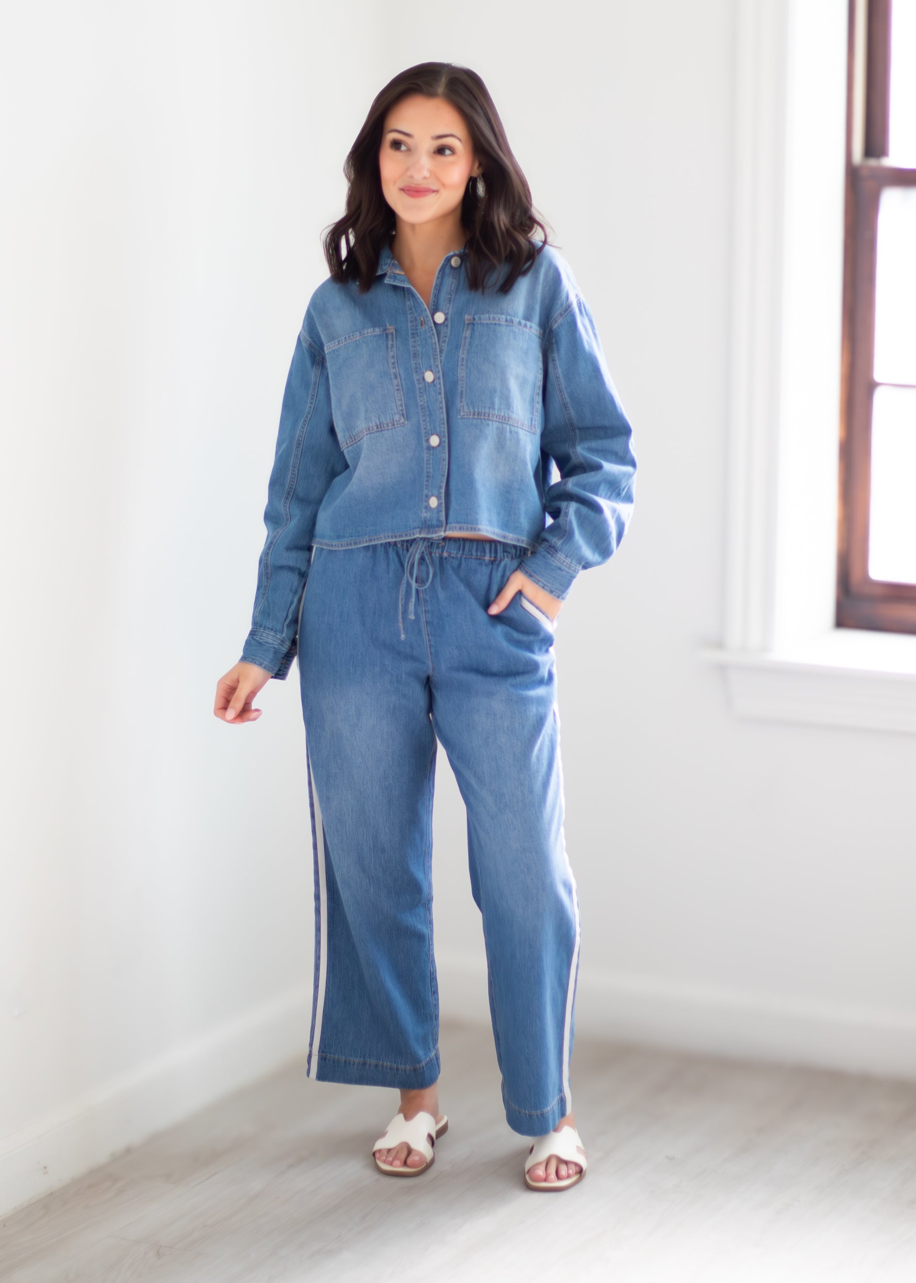 A woman modeling a matching blue denim set consisting of a cropped button-up shirt and wide-leg drawstring pants with side stripes, standing in a bright white room.