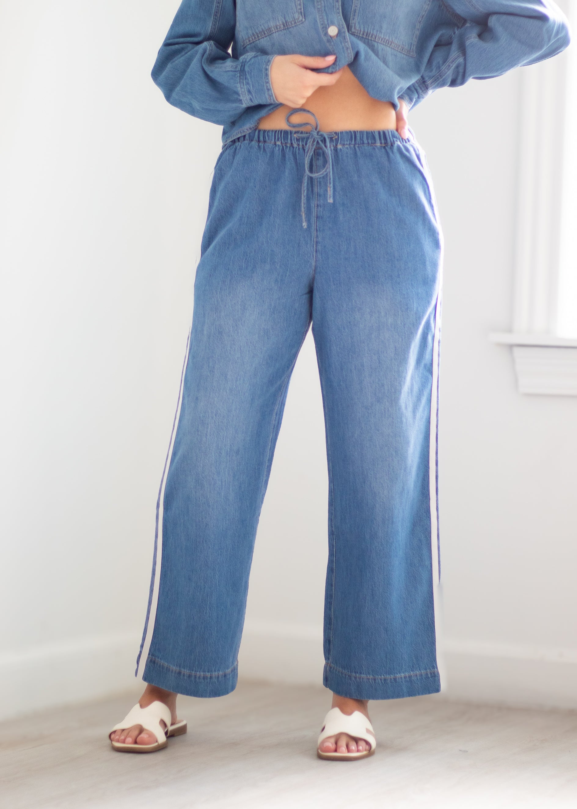 A person wearing wide-leg blue denim pants with white side stripes, paired with a cropped blue denim shirt and white slide sandals, standing in a bright indoor room.