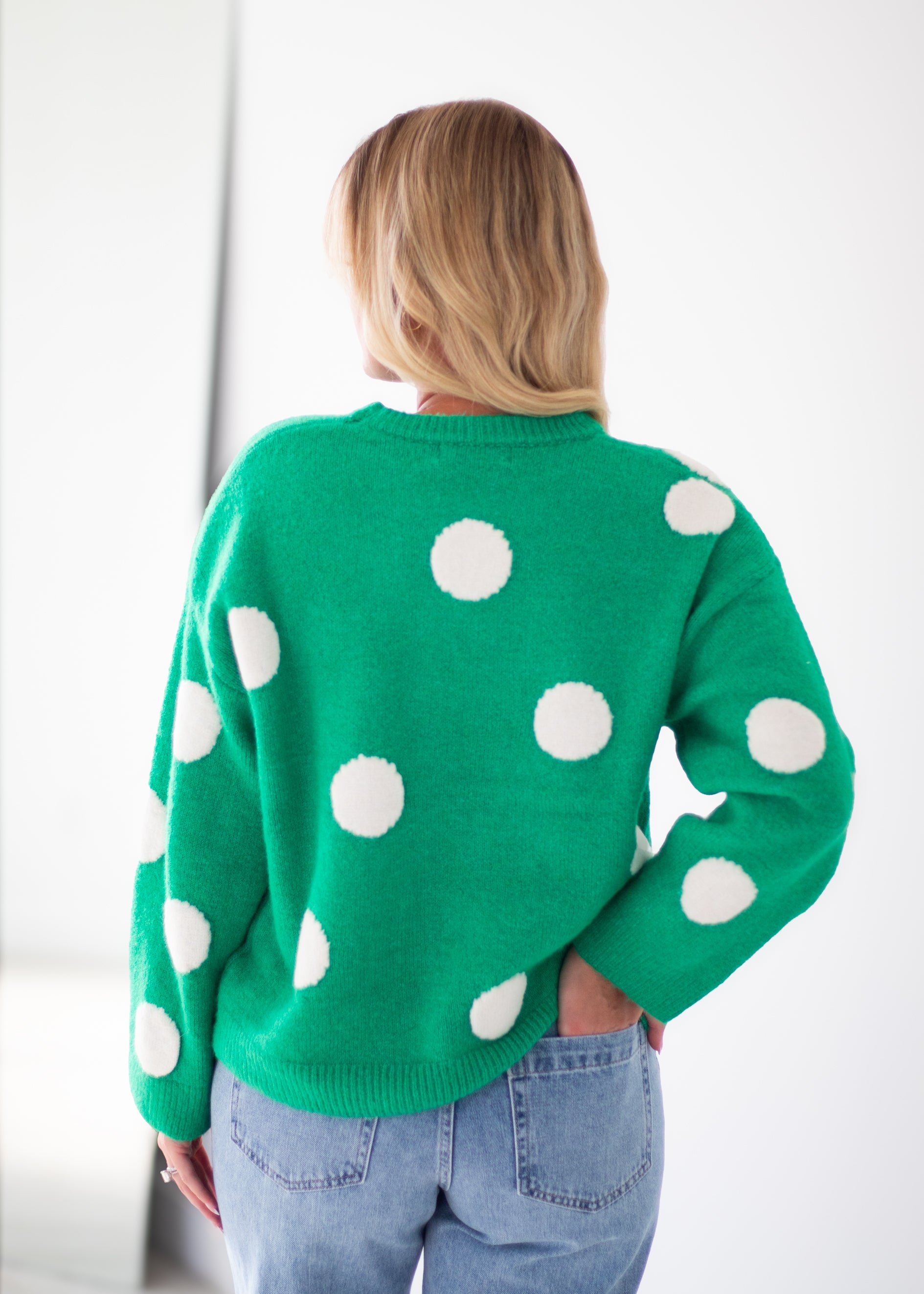 A woman viewed from behind wearing a bright green knit sweater with large white polka dots, paired with light blue jeans, against a plain white background.