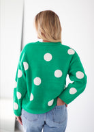 A woman viewed from behind wearing a bright green knit sweater with large white polka dots, paired with light blue jeans, against a plain white background.