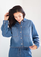 A woman wearing a blue denim long-sleeve button-up shirt and matching denim drawstring pants, posing against a light gray studio background.