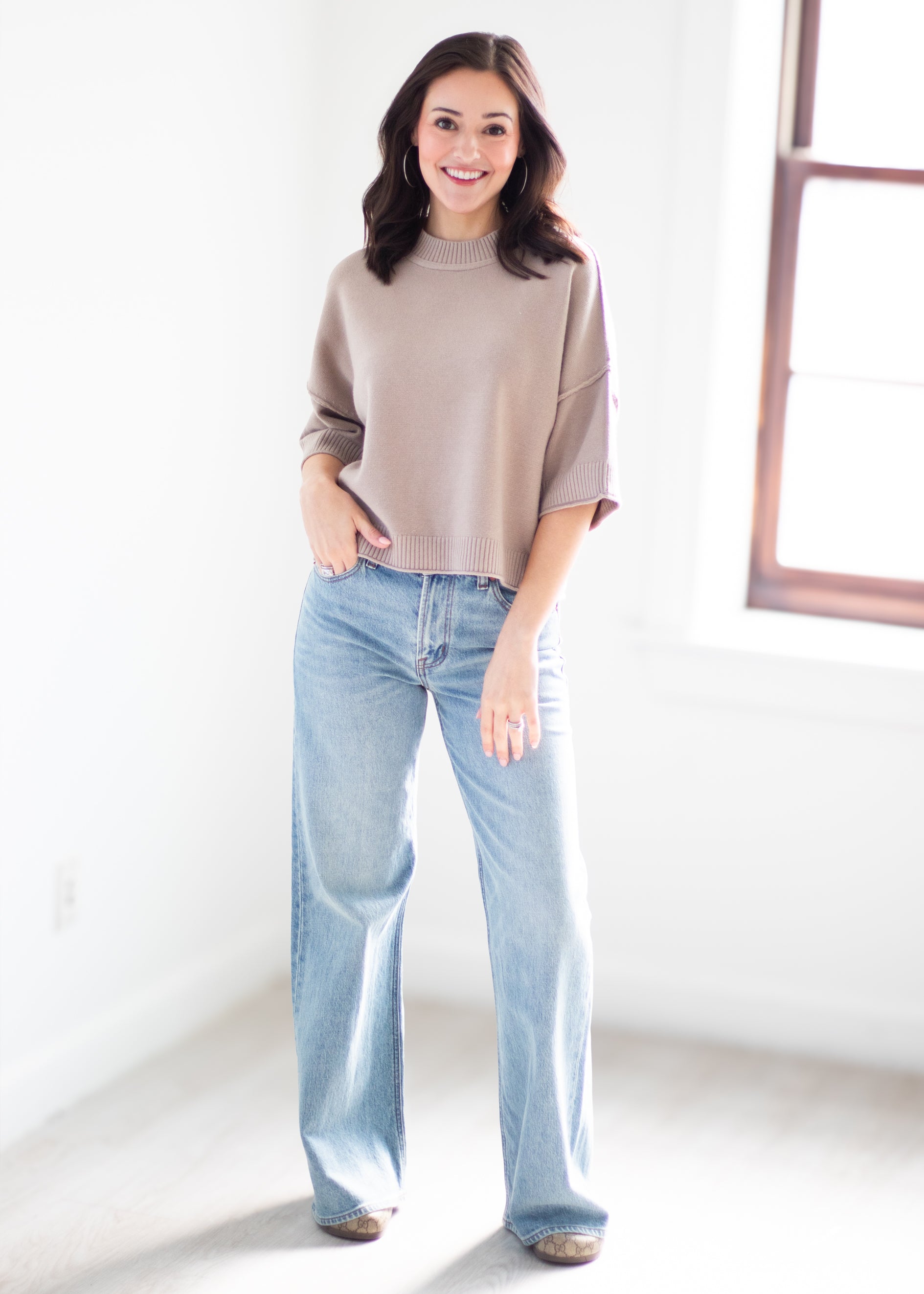 A woman stands in a bright, minimalist room wearing a taupe knit sweater and light-wash wide-leg jeans.