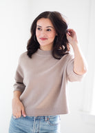 A woman with dark brown hair wearing a light taupe knit mock neck sweater and blue jeans, posing against a bright white background.