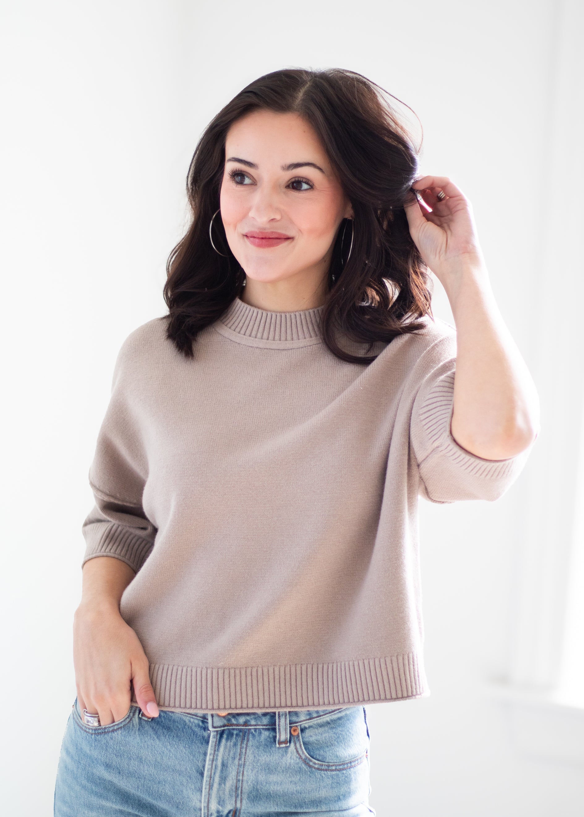 A woman with dark brown hair wearing a light taupe knit mock neck sweater and blue jeans, posing against a bright white background.