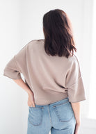 A woman viewed from behind wearing a light taupe knit sweater with elbow-length sleeves and light blue jeans, set against a bright white background.