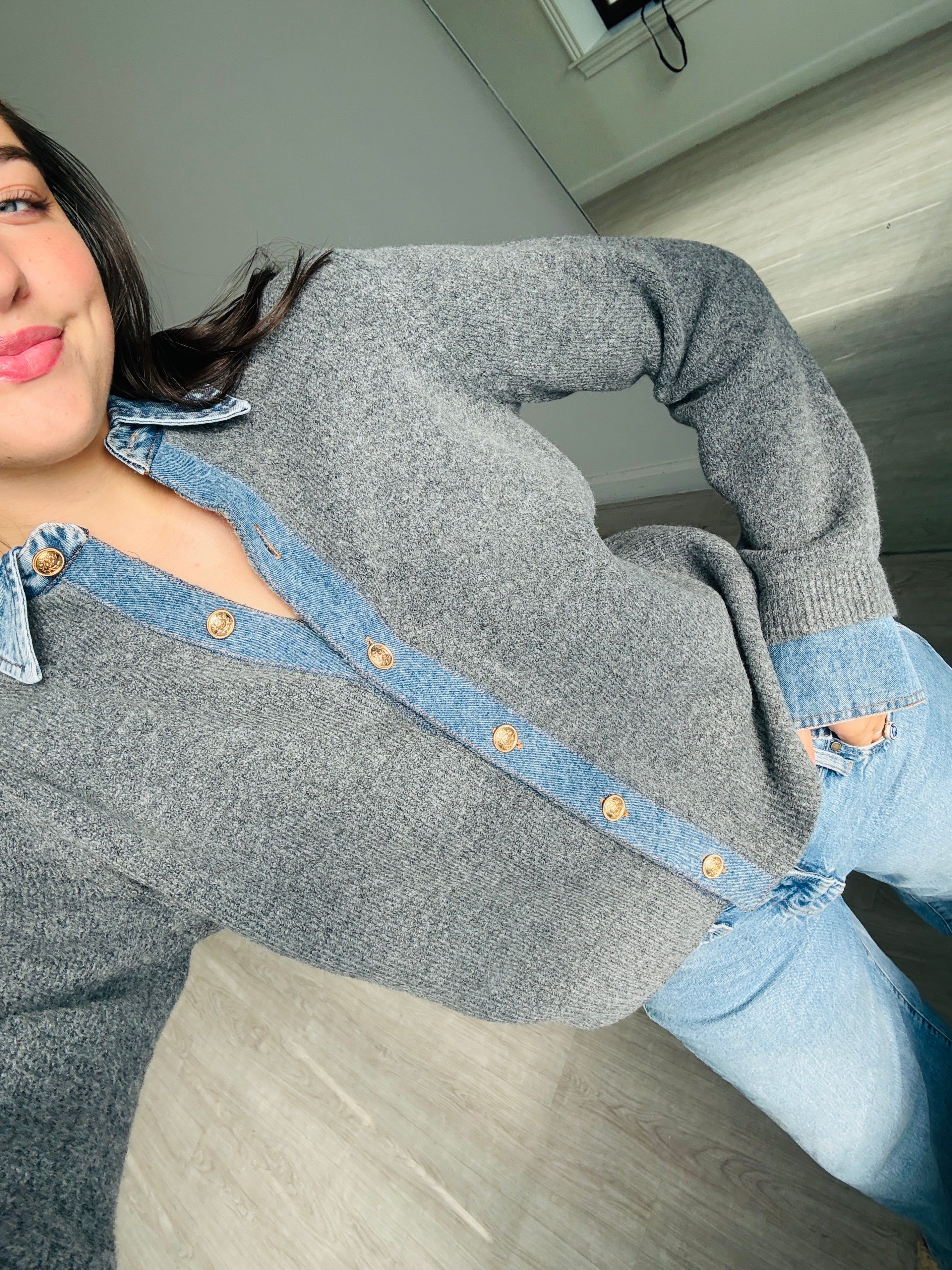 A person wearing a gray knit cardigan with a light blue denim collar and button placket, paired with light blue jeans, photographed indoors on a wood floor.