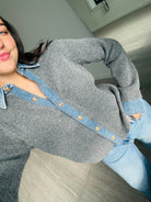 A person wearing a gray knit cardigan with a light blue denim collar and button placket, paired with light blue jeans, photographed indoors on a wood floor.