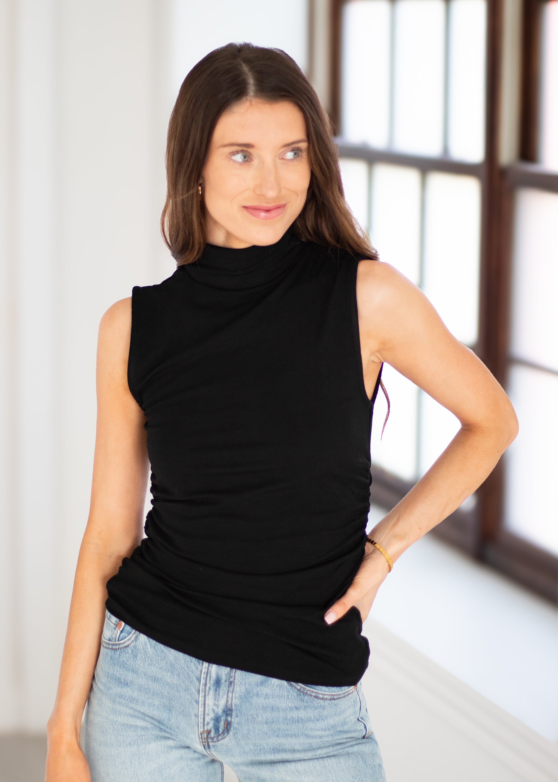 A woman wearing a sleeveless black mock neck top and light wash blue jeans, standing in a brightly lit room with a window in the background.