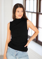 A woman wearing a sleeveless black mock neck top and light wash blue jeans, standing in a brightly lit room with a window in the background.