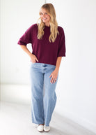 A woman modeling an oversized short-sleeve crewneck top in deep burgundy, paired with light-wash wide-leg jeans and white shoes, standing against a bright white wall.