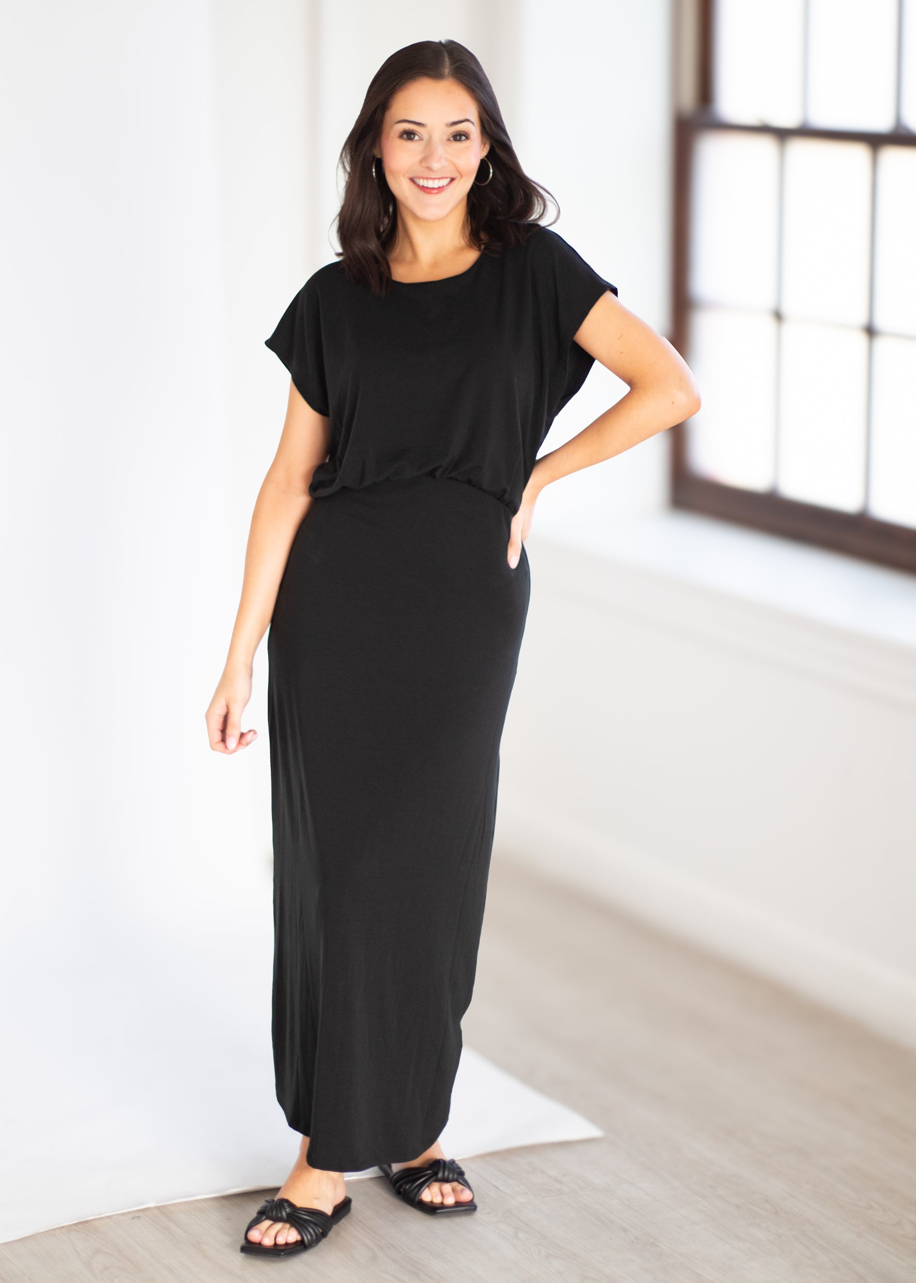 A woman modeling a black short-sleeve maxi dress with side pockets, paired with black slide sandals, standing in a bright white room with a window.