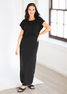 A woman modeling a black short-sleeve maxi dress with side pockets, paired with black slide sandals, standing in a bright white room with a window.