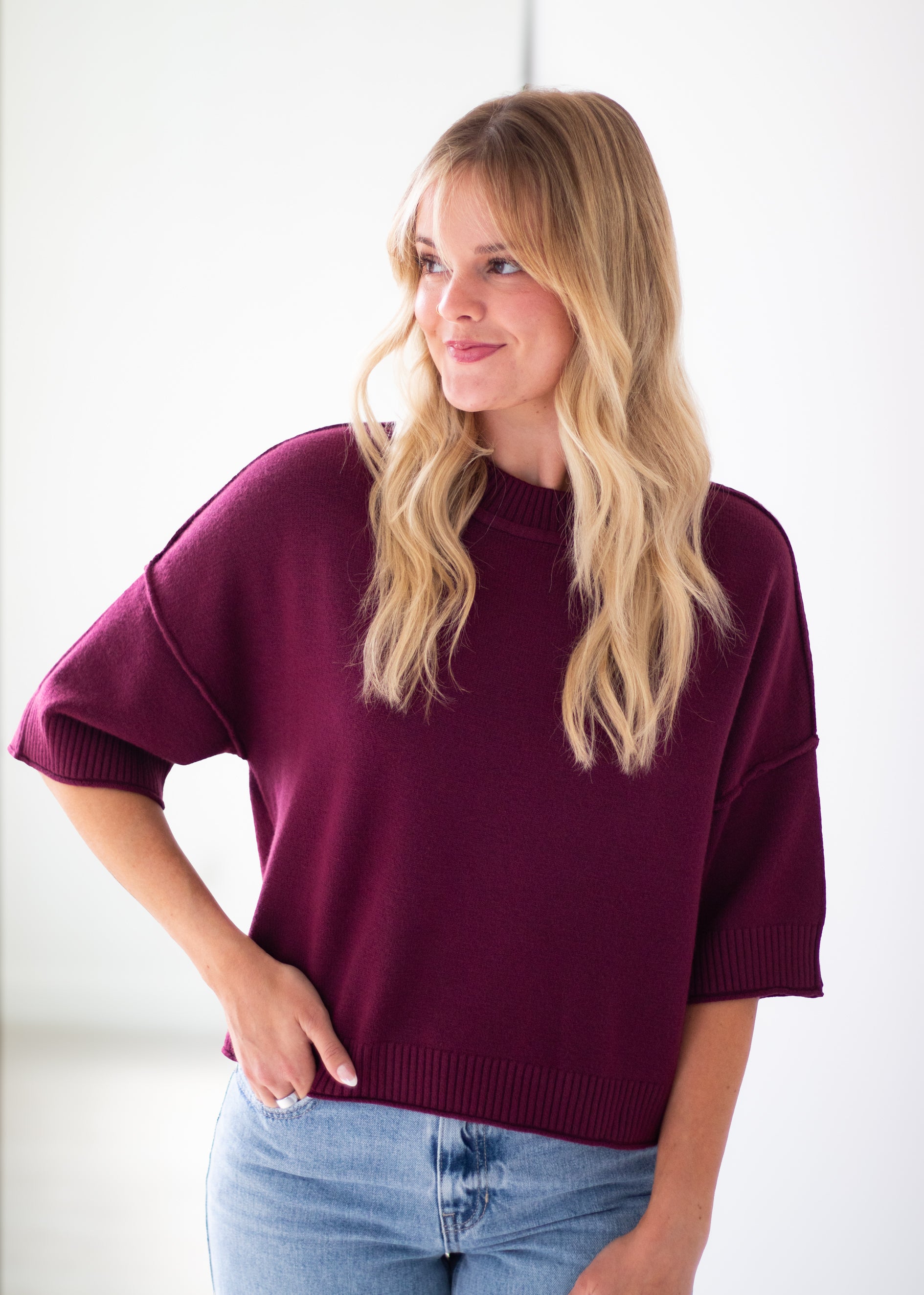 A woman with blonde hair wearing an oversized short-sleeve knit sweater in deep burgundy and light blue jeans, standing against a plain white background.