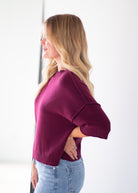 A woman with blonde hair is shown in profile wearing a short-sleeve knit sweater in deep burgundy and light blue jeans against a bright white background.