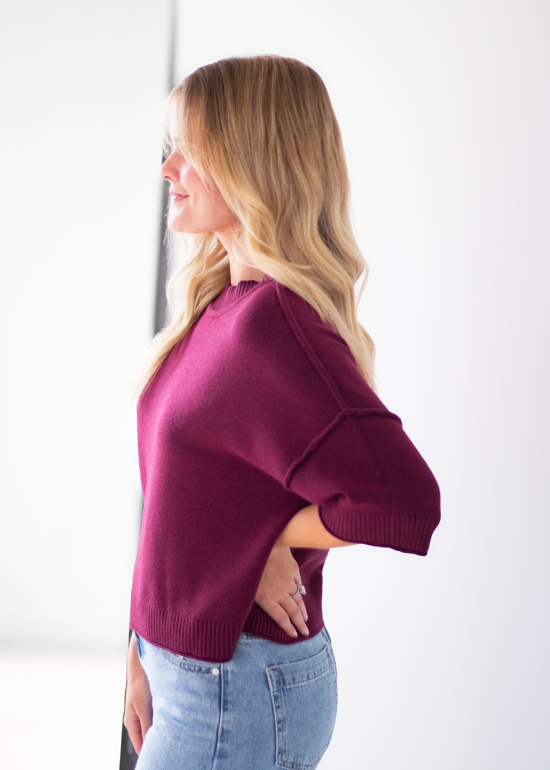 A woman with blonde hair is shown in profile wearing a short-sleeve knit sweater in deep burgundy and light blue jeans against a bright white background.