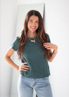 A woman with long brown hair wearing a dark green short-sleeve t-shirt and light blue jeans, standing in a brightly lit room.