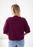 A woman viewed from behind wearing a short-sleeve knit sweater in deep burgundy and light blue jeans against a plain white background.