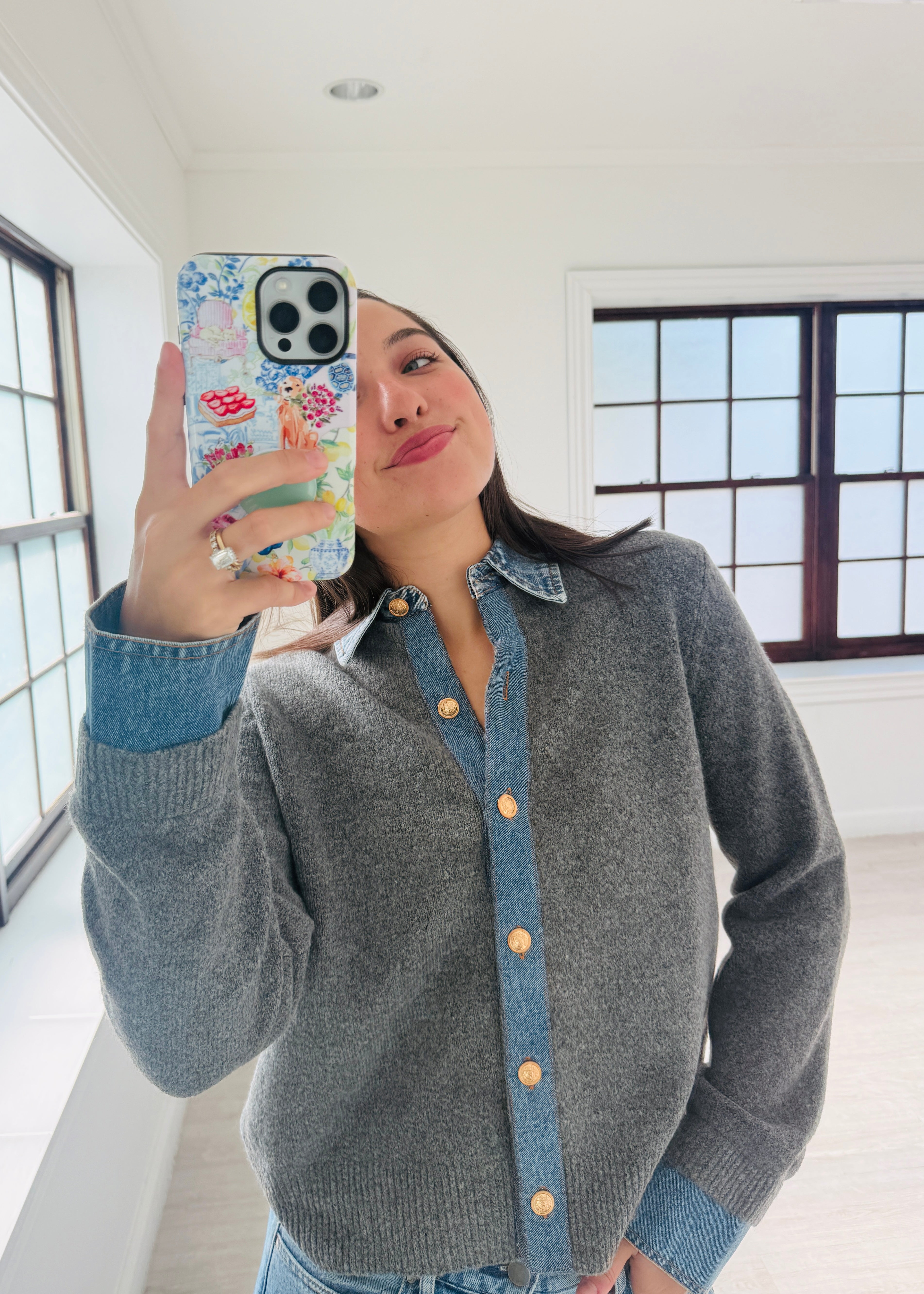 A woman takes a mirror selfie wearing a gray knit button-up cardigan with a denim collar and cuffs, paired with a denim shirt underneath. She is holding a floral-patterned phone case in a bright, modern room with large windows.