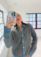 A woman takes a mirror selfie wearing a gray knit button-up cardigan with a denim collar and cuffs, paired with a denim shirt underneath. She is holding a floral-patterned phone case in a bright, modern room with large windows.