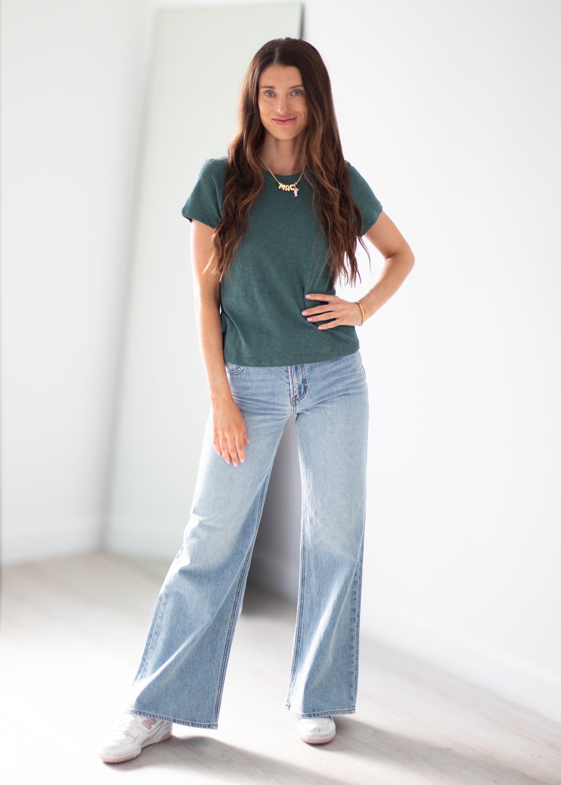 A woman modeling a dark green short-sleeve knit t-shirt and light-wash wide-leg denim jeans, standing in a bright, minimalist white room.