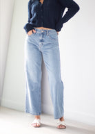 A person wearing light-wash wide-leg denim jeans, a dark navy ribbed long-sleeve top, and white slide sandals, standing against a plain white wall.