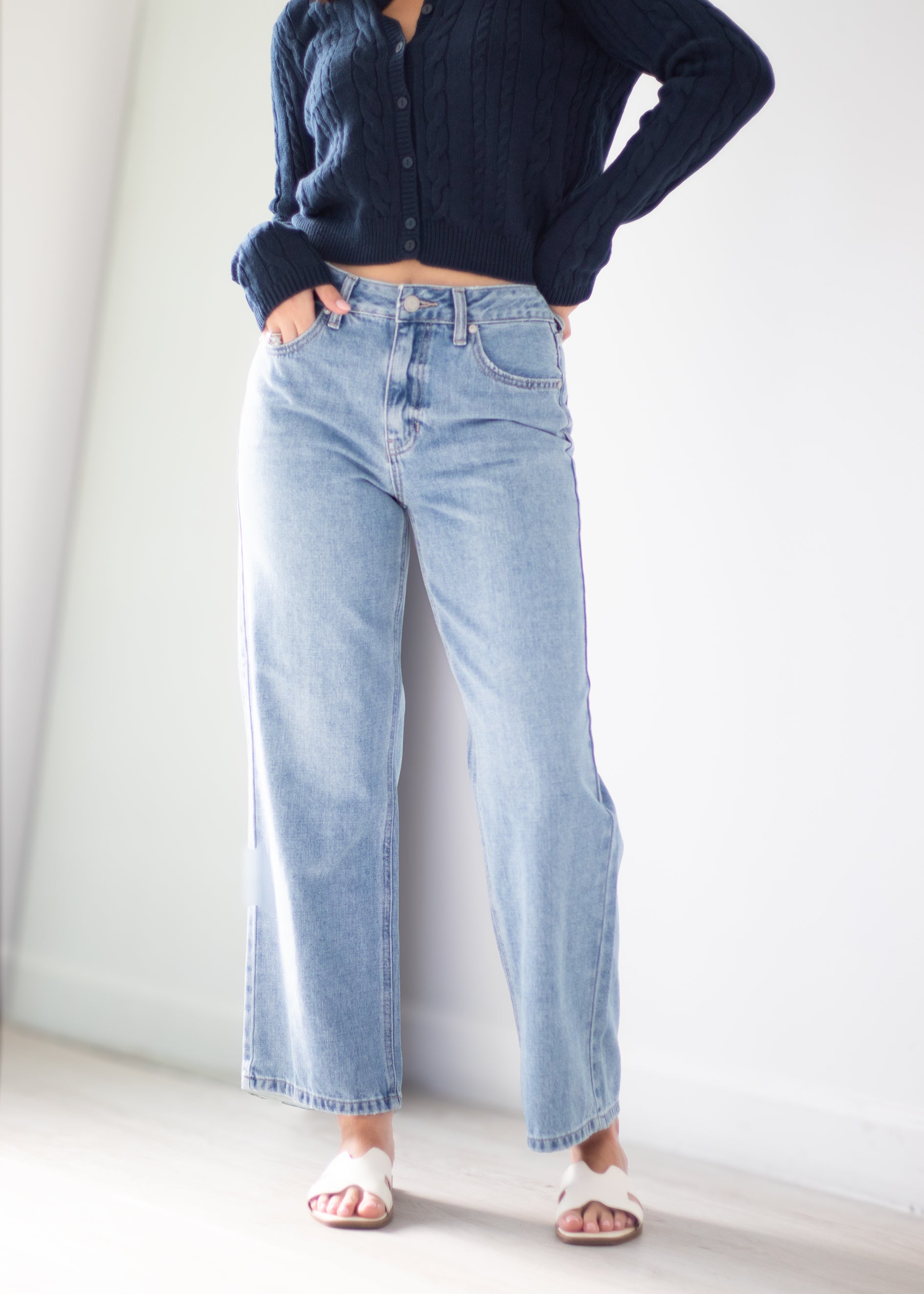 A person wearing light-wash wide-leg denim jeans, a dark navy ribbed long-sleeve top, and white slide sandals, standing against a plain white wall.