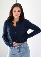 A woman wearing a navy blue cable-knit button-up cardigan sweater and light-wash blue jeans, posing against a clean white studio background.