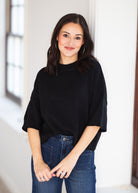 A woman with dark hair wearing a black knit short-sleeve top and blue jeans, standing in a brightly lit indoor setting.