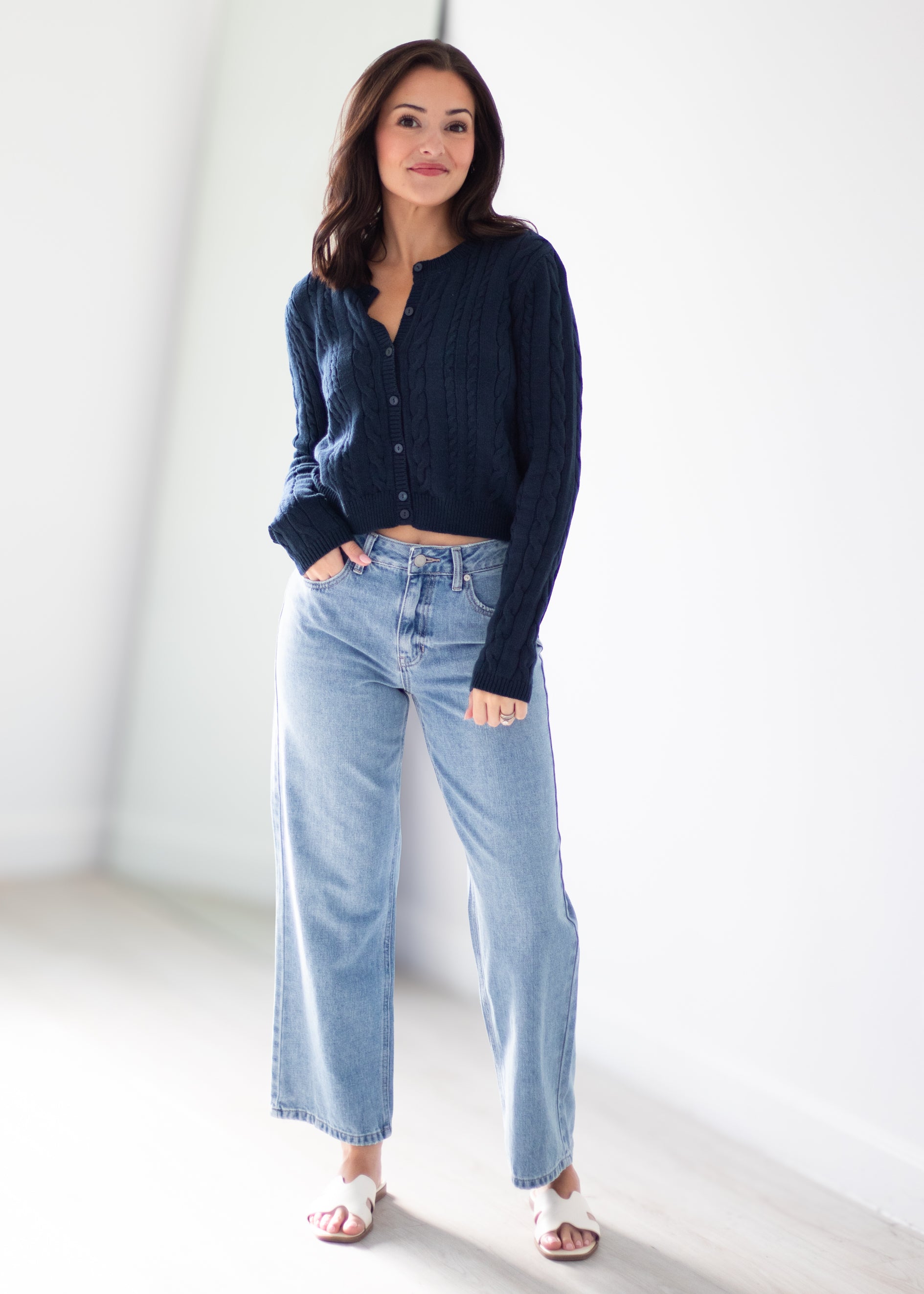 A woman modeling a dark navy blue ribbed knit button-up cardigan and light-wash wide-leg blue jeans, standing in a bright, minimalist white room.