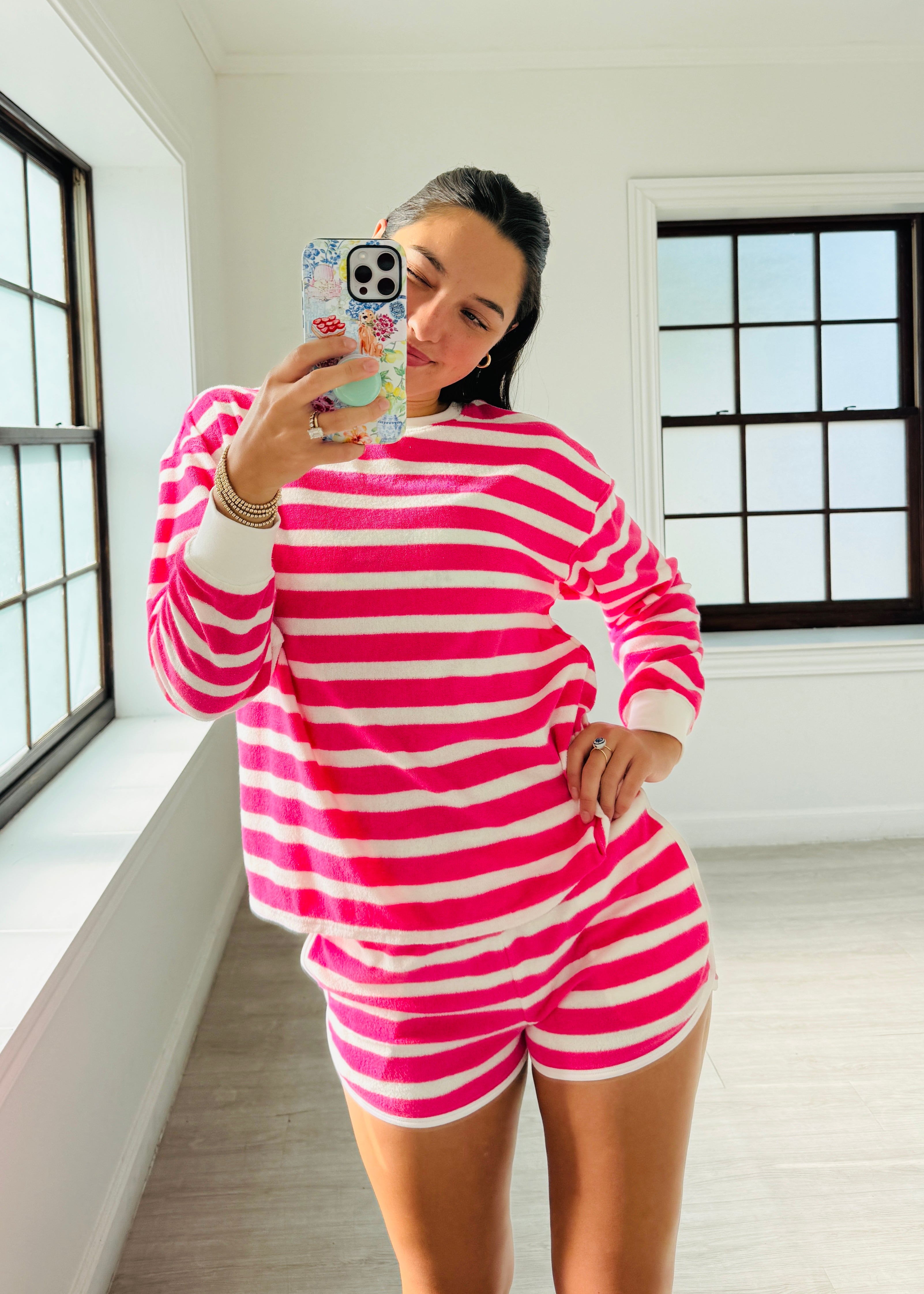 A woman takes a mirror selfie wearing a matching pink and white striped lounge set consisting of a long-sleeve top and shorts, in a bright room with large black-framed windows.