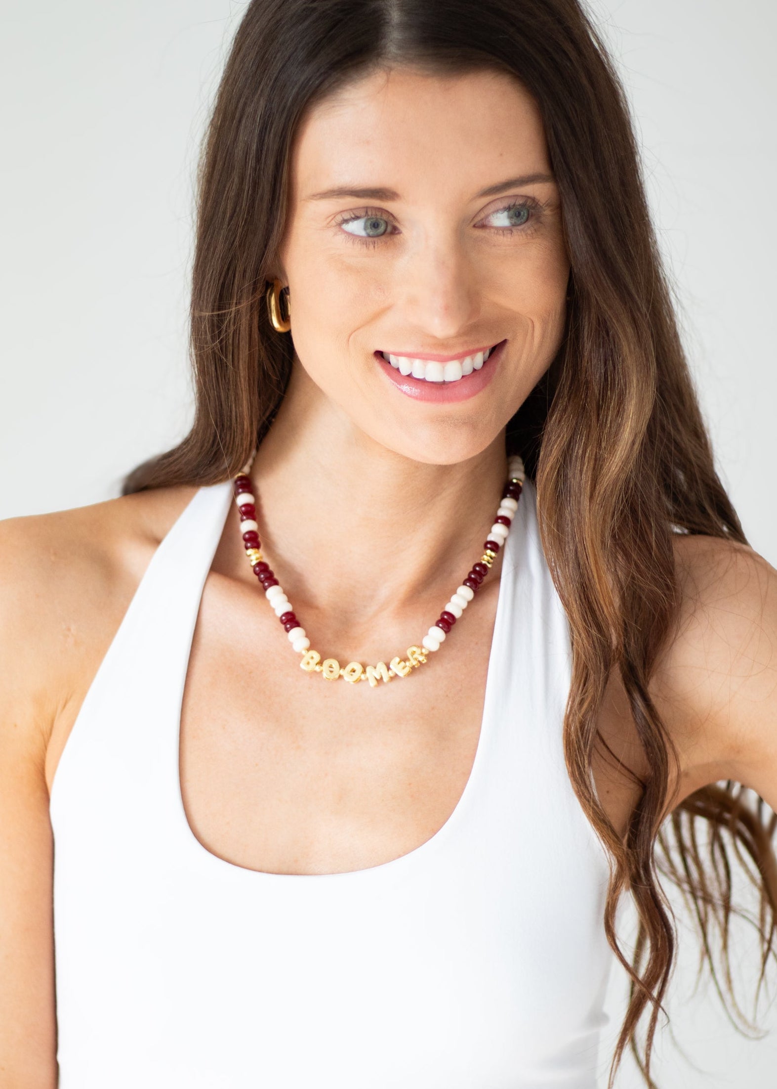 A woman with long brown hair wearing a white tank top and a beaded necklace with red, white, and gold beads, photographed against a light gray background.