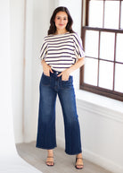 A woman modeling a white short-sleeve top with thin navy horizontal stripes and dark blue wide-leg cropped jeans, standing in a bright room with a window.
