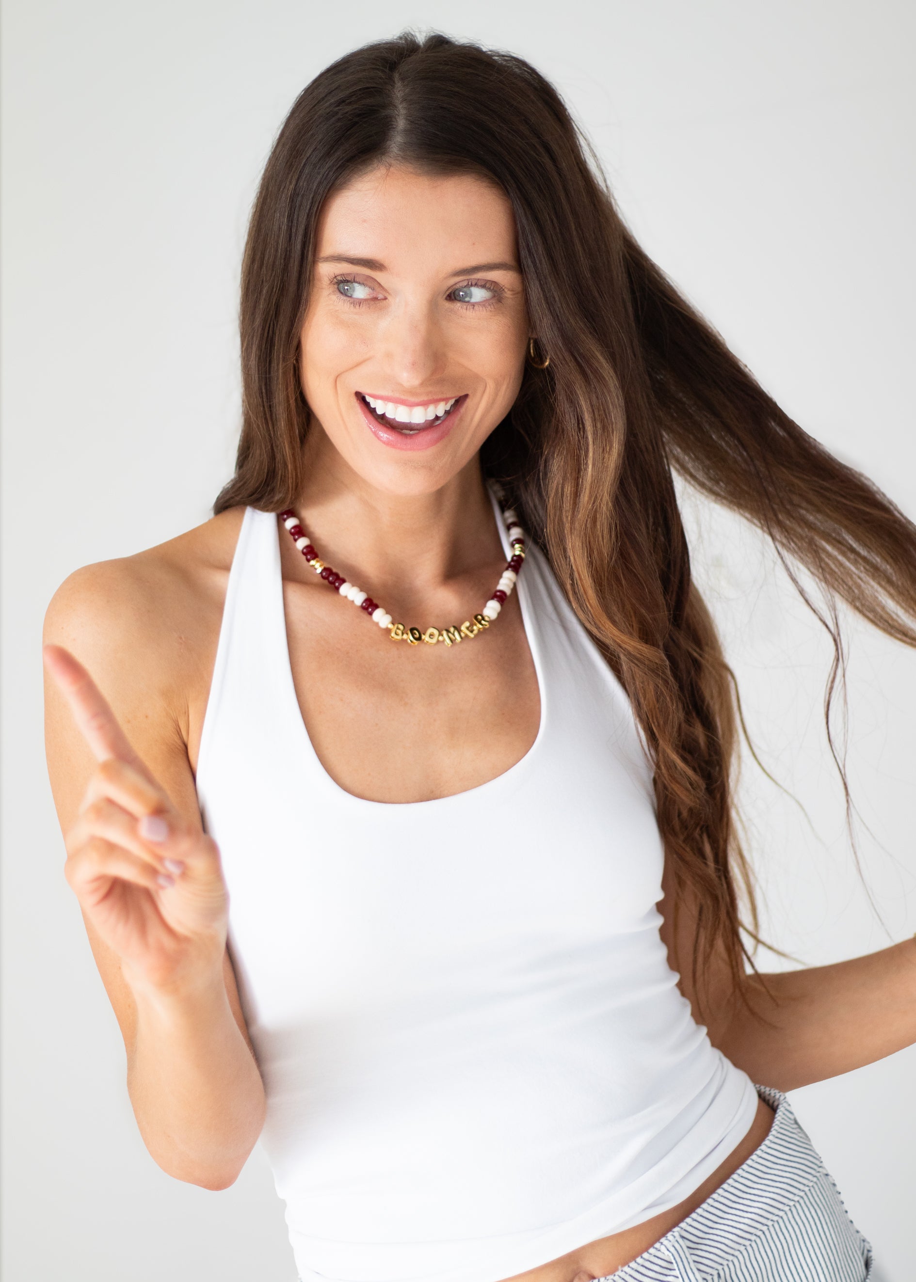 A smiling woman with long brown hair wearing a white tank top and a multi-colored beaded necklace, pointing towards the viewer against a light gray background.