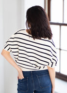 A woman viewed from behind wearing a cream-colored knit top with black horizontal stripes and blue jeans, standing in a brightly lit room with a window.