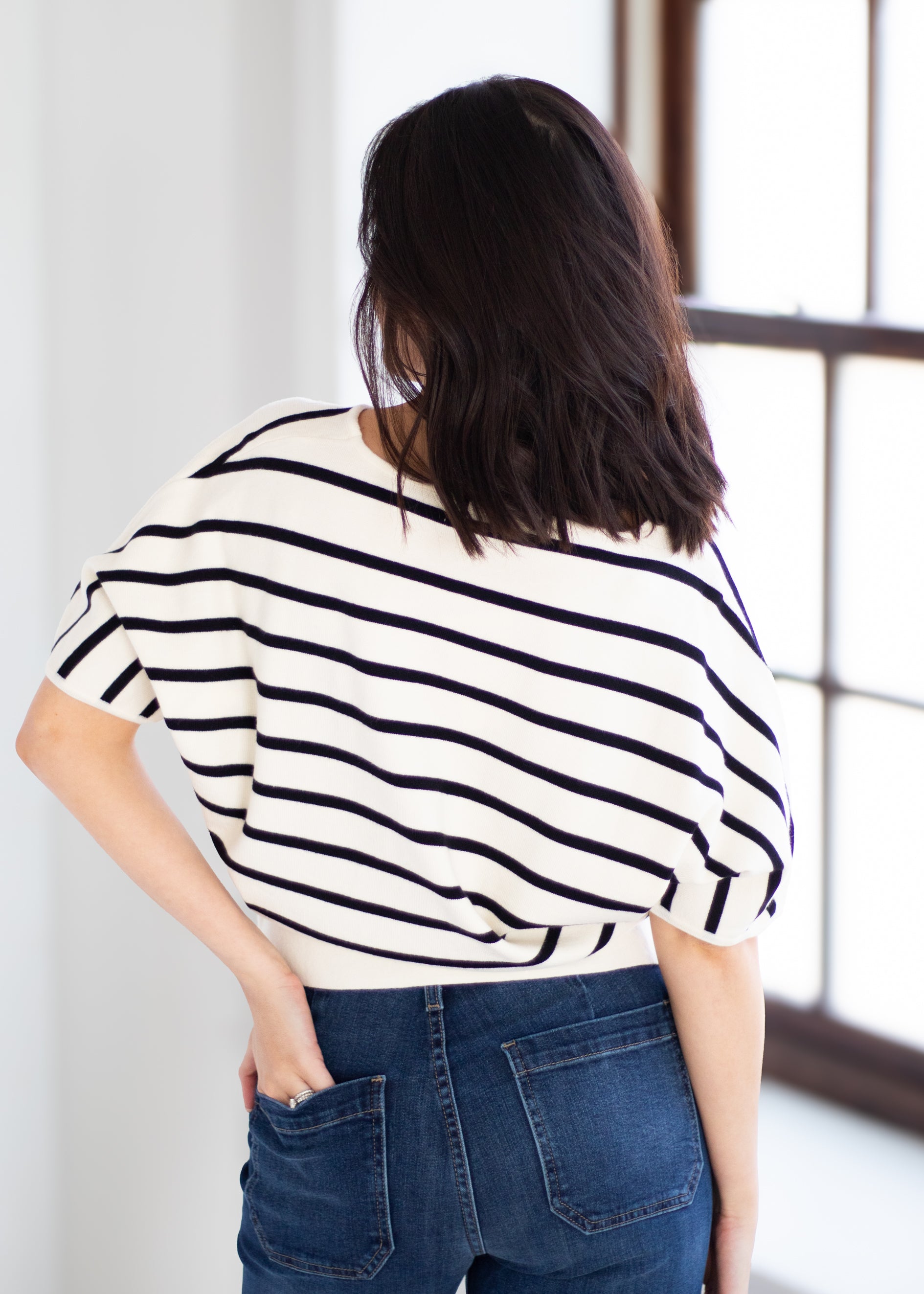 A woman viewed from behind wearing a cream-colored knit top with black horizontal stripes and blue jeans, standing in a brightly lit room with a window.