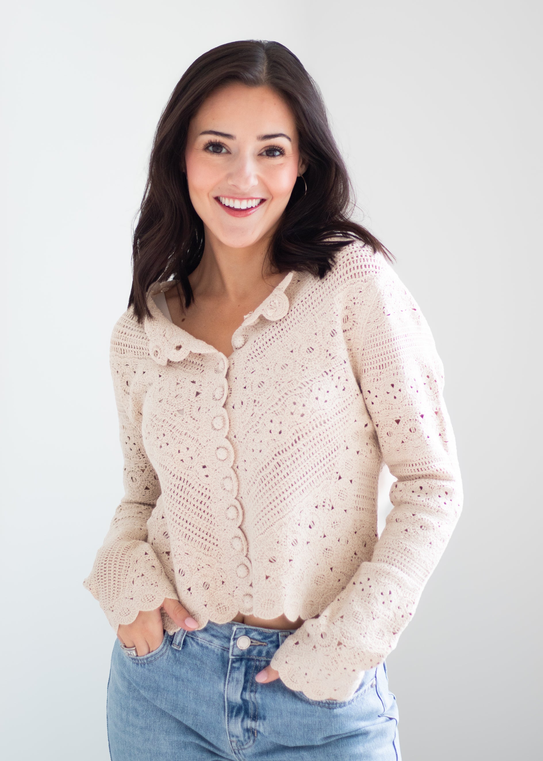 A smiling woman with dark hair wearing a light beige eyelet knit cardigan with scalloped edges and button details, paired with blue jeans, against a plain light gray background.